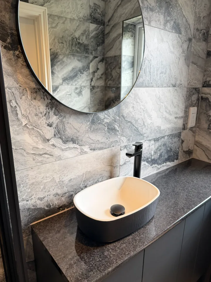 Bathroom with marble-effect tiling, black vanity unit and round black mirror by CMS Plumbing & Bathrooms